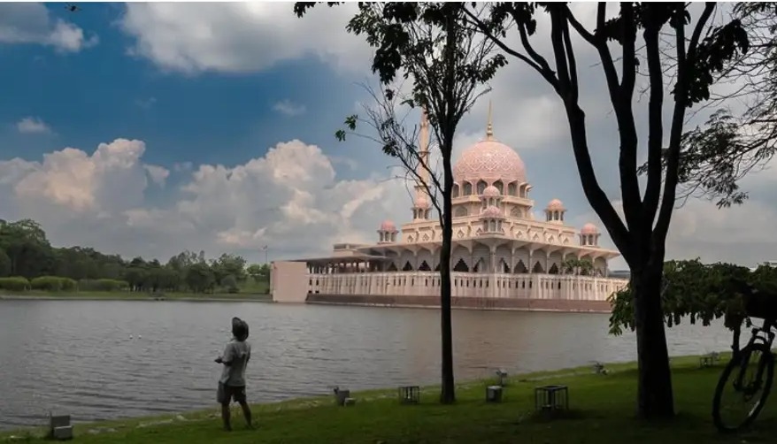 Pink-domed Putra Mosque across the lake, framed by trees, with a boy on the grass
