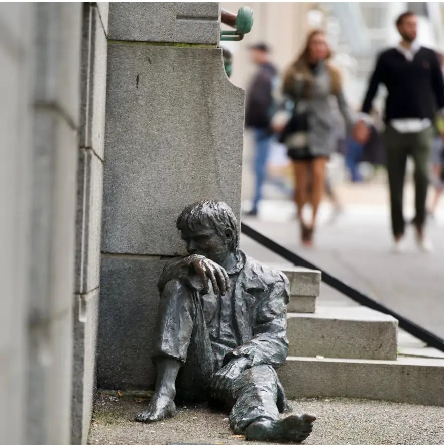 Bronze statue of a resting boy against a stone wall as pedestrians pass