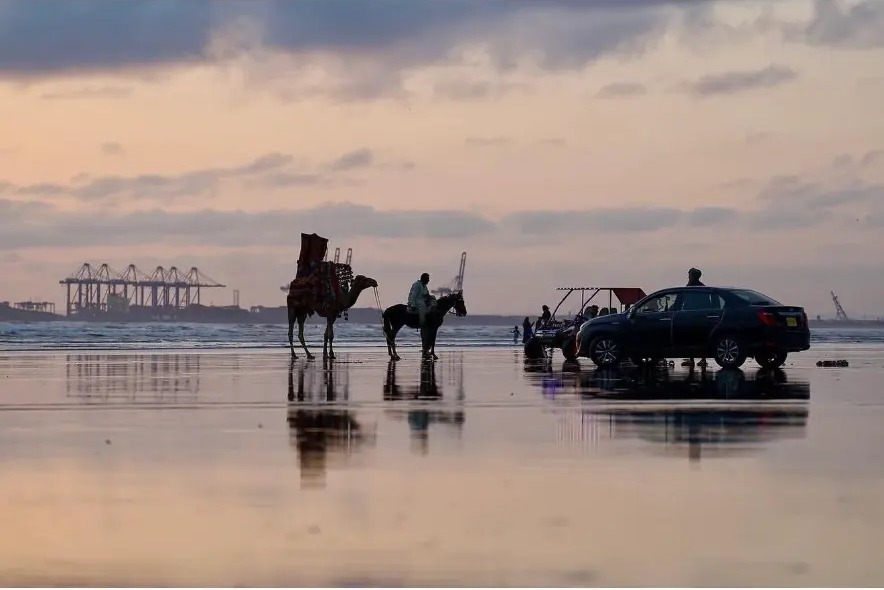 Camel and horse riders reflected on the wet sand at sunset on Sea View Beach, Karachi, Pakistan