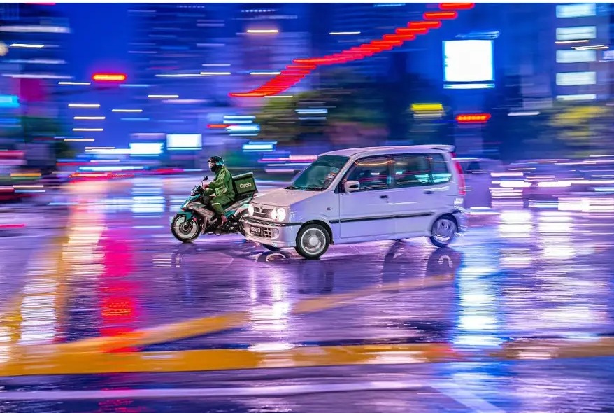 Panning shot of a delivery rider and small car in neon night traffic