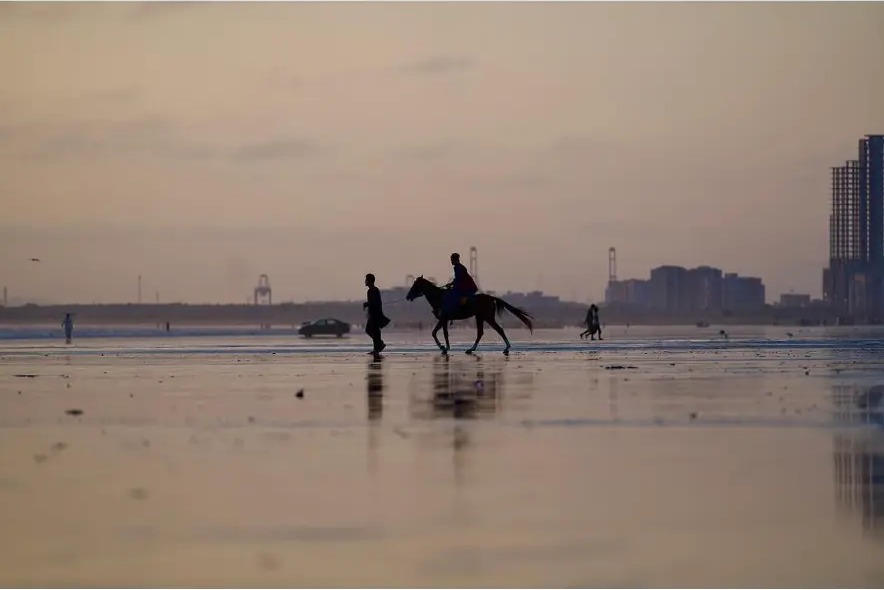 Horse rider crossing the wet sand with the city skyline behind in Karachi, Pakistan
