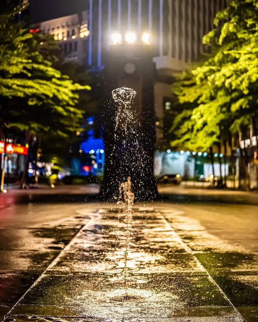 Street fountain lit at night with wet paving stretching away