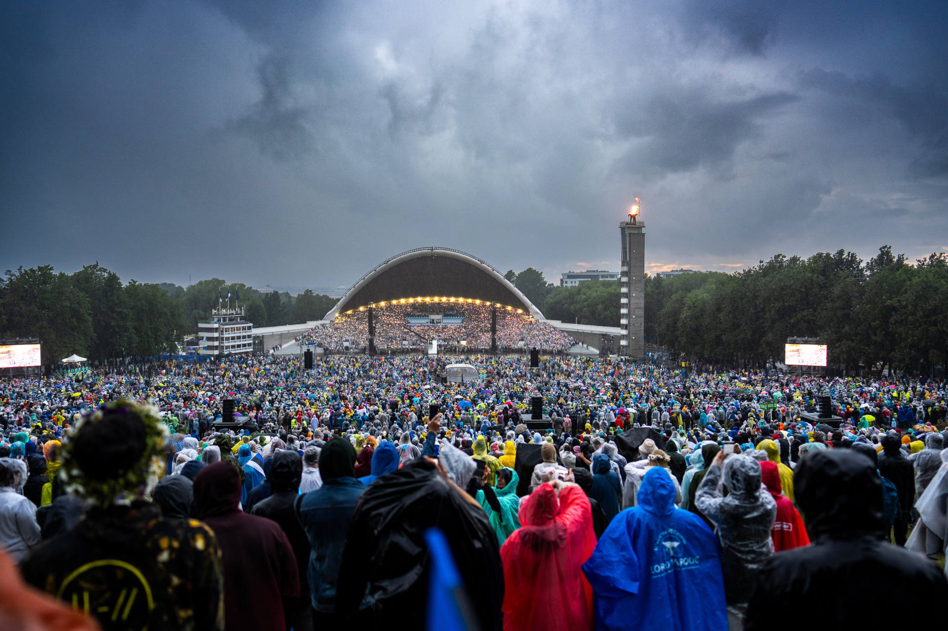 Huge crowd in rain ponchos facing the Song Festival Grounds stage under storm clouds, Tallinn, Estonia