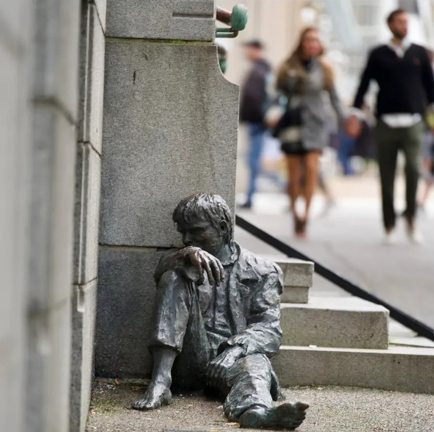 Bronze statue of a resting boy against a stone wall as pedestrians pass