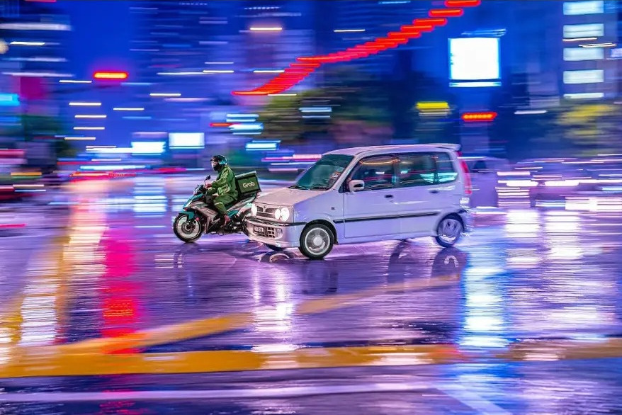 Panning shot of a delivery rider and small car in neon night traffic