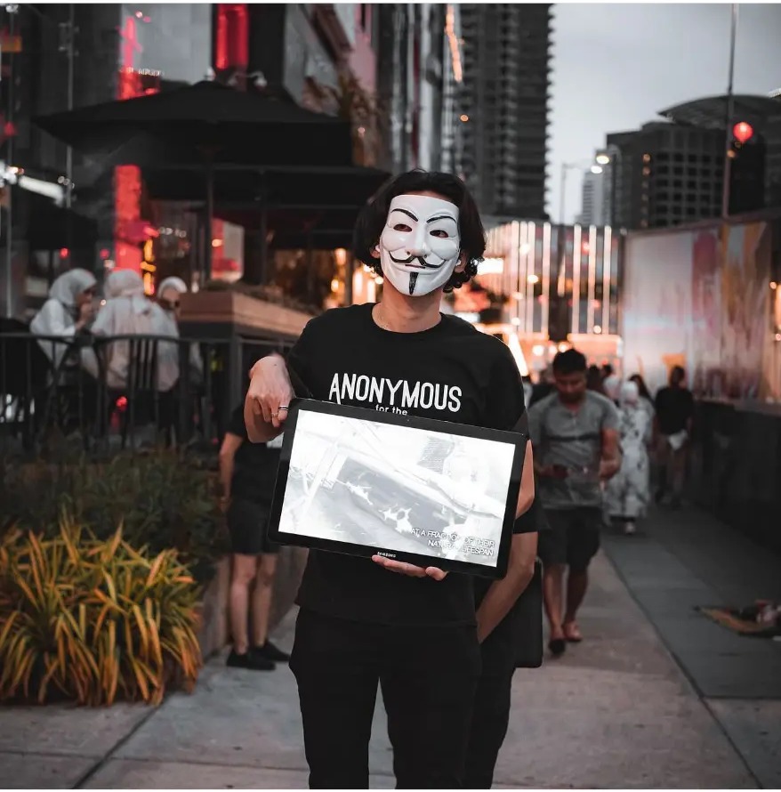 Activist in an Anonymous mask holding a screen on a busy street