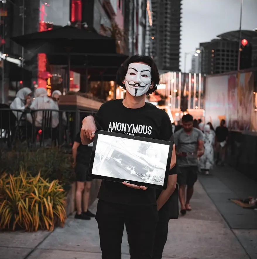 Activist in an Anonymous mask holding a screen on a busy street