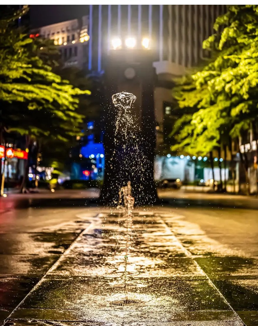 Street fountain lit at night with wet paving stretching away